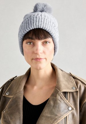 Szary, dziany beanie z pomponem na czubku, prezentujący prążkowaną teksturę. Noszony z brązową skórzaną kurtką na czarnym swetrze.