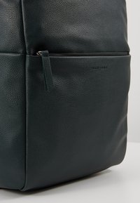 Gros plan sur un sac en cuir texturé foncé avec une poche zippée horizontale et une tirette de fermeture éclair en cuir.