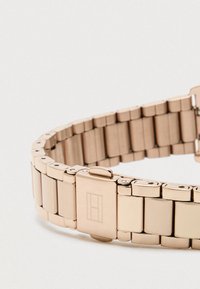 Náramek na hodinky z růžově zlatého kovu s propojeným designem a sponou, která má gravírované obdélníkové logo na světlém pozadí.