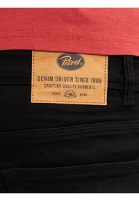 Czarne dżinsy z denimu, z beżową skórzaną metką oraz niebieskim tekstem. Metka głosi "Petrol" oraz "Denim Driven Since 1989."
