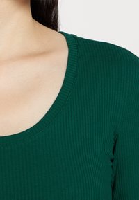 Grön ribbad topp med vid rundad halsringning och strukturerad tyg, som framhäver dess figurnära passform och släta yta.