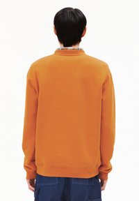 Persona in piedi di spalle che indossa una felpa arancione e pantaloni cargo blu su uno sfondo bianco semplice.