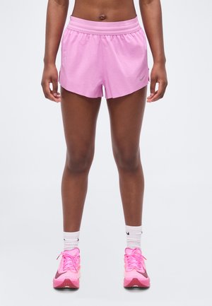 Sportowiec stoi, ubrany w jasnoróżowe spodenki, białe skarpetki Nike oraz jaskrawo różowe buty do biegania na jednolitym jasnym tle.