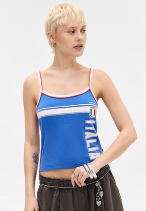 Jeune femme aux cheveux blonds courts portant un débardeur bleu de l’Italie et un pantalon foncé, posant avec la main près de la taille et regardant sur le côté.