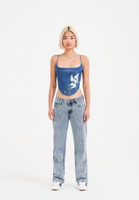 Top corto in denim con ricami floreali; abbinato a jeans azzurri chiari con motivi ricamati bianchi; modello che indossa sneakers bianche.