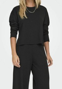 Schwarzer gerippter Strickpullover mit langen Ärmeln und cropped Saum. Weicher Stoff und lockere Passform. Getragen mit passenden schwarzen gerippten Hosen.