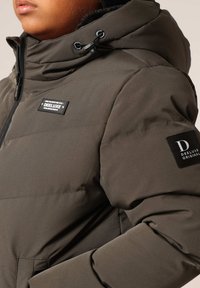 Veste matelassée verte olive avec capuche, dotée d'une fermeture éclair à l'avant, d'un tissu texturé et de patchs de marque sur la poitrine et la manche.