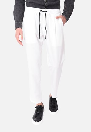 Uomo che cammina con pantaloni bianchi con coulisse, camicia di denim nera e sneakers nere con suola bianca su uno sfondo semplice.