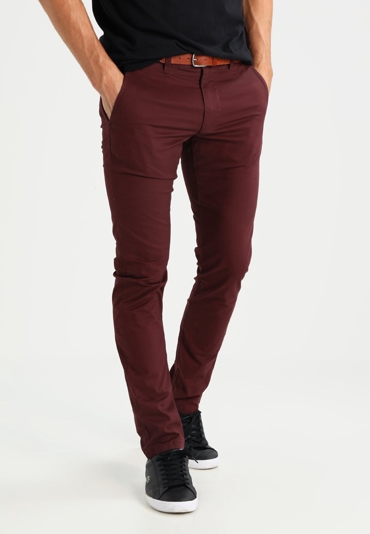 Burgundy slim-fit byxor med en slät textur och bältesöglor, kombinerat med svarta lågt skurna sneakers, händerna i fickorna, stående pose.