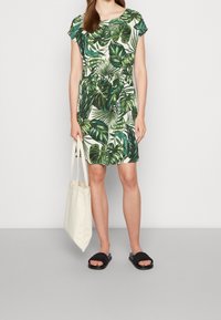 Šaty s krátkým rukávem s zeleným tropickým listovým vzorem na světlém pozadí, elastický pas, kombinované s béžovou taškou a černými sandály.