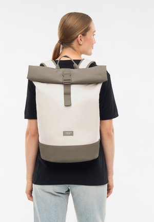 Femme portant un sac à dos à enroulement beige et gris par-dessus une chemise noire et un jean bleu clair, vue de dos sur fond blanc.