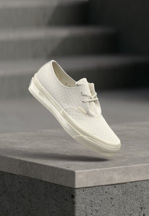 White low-top sneaker with a subtle checkerboard pattern, white laces, and a thick sole resting on a gray concrete ledge.