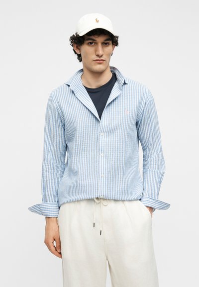 Νέος άνδρας που φοράει λευκό καπέλο, φωτεινό γαλάζιο ριγέ πουκάμισο με κουμπιά πάνω από μαύρο μπλουζάκι και λευκό παντελόνι με κορδόνι, με τα χέρια εν μέρει στις τσέπες.