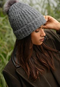 Šedá pletená čepice s kožíškem na vrchu, s texturovaným vzorem a ohrnutým lemem, nošená s tmavým pláštěm.