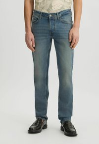 Jeans de mezclilla azules con corte recto, acabado desgastado, bolsillos frontales y cierre de botón, combinados con mocasines negros.