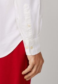 Mano descansando al lado, vistiendo una camisa blanca de botones en la muñeca y pantalones rojos, contra un fondo liso de color gris claro.