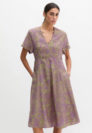 Femme aux cheveux foncés attachés en queue de cheval basse, portant une robe midi vert olive avec des manches courtes retroussées et un motif floral violet, mains dans les poches.