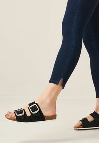 Czarne zamszowe klapki na płaskiej podeszwie z dwoma dużymi kwadratowymi klamrami i korkowym wkładką, w zestawieniu z ciemnoniebieskimi dopasowanymi legginsami z rozcięciami na kostkach.