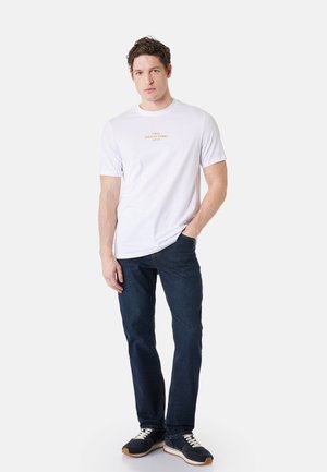 Mand står med den ene hånd i lommen iført en hvid t-shirt med gul tekst "Italy Amalfi Coast Naples", mørke jeans og sorte sneakers.