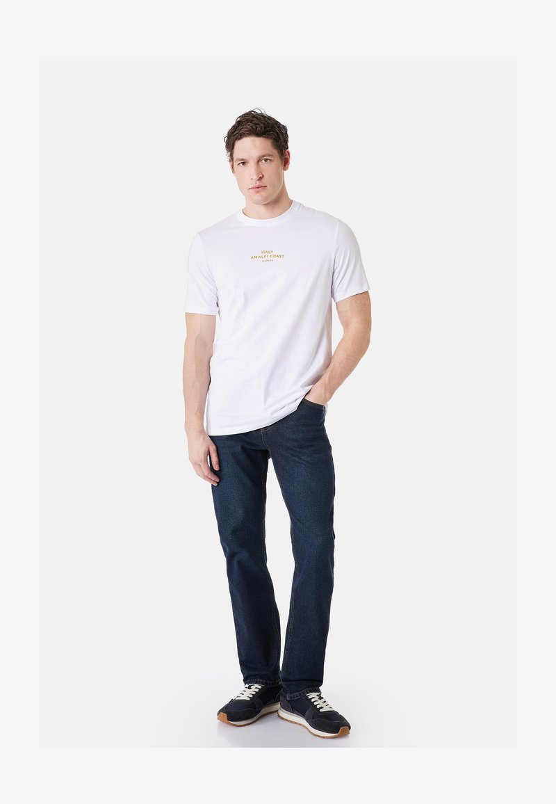 Homme debout, une main dans la poche, portant un t-shirt blanc avec le texte jaune « Italy Amalfi Coast Naples », jean foncé et baskets noires.