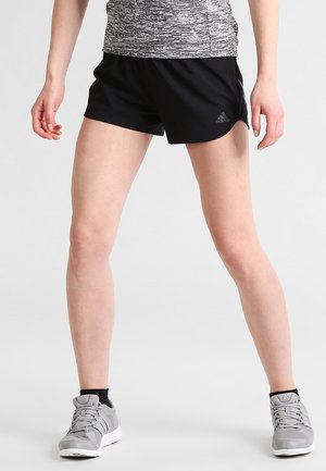 Person står med benene fra hinanden iført sorte Adidas-shorts, grå løbesko og en grå mønstret top mod en ensfarvet lys baggrund.