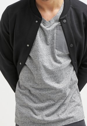 Personne portant un T-shirt gris à col en V avec une poche poitrine sombre et une veste noire boutonnée par-dessus.