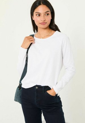 Langärmliges, weißes Hemd mit rundem Ausschnitt, kombiniert mit dunkelblauen Jeans. Das Hemd hat eine lockere Passform und besteht aus einem glatten Stoff.