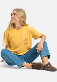Gul bomull T-skjorte med en liten brun grafikk på venstre bryst, kombinert med blå denimjeans og brune slip-on sko.