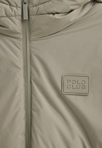 Giubbotto imbottito color kaki con una texture liscia, dotato di cerniera completa e di un'etichetta rettangolare 'POLO CLUB' ricamata sul petto.