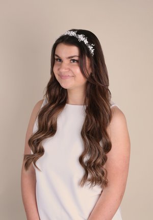 Weiße Blumenhaube mit kleinen Blüten und Blättern, getragen von einer Person mit langen, gewellten braunen Haaren, vor einem neutralen Hintergrund.