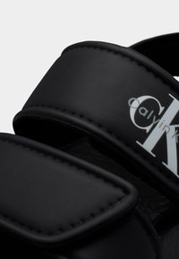 Μαύρο σανδάλι με φαρδιές λωρίδες, που φέρει λευκό λογότυπο Calvin Klein στη επάνω λωρίδα, κοντινή όψη.