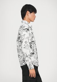 Camicia a maniche lunghe in tessuto bianco con ornate fantasie floreali nere, con colletto a bottoni e una vestibilità comoda. Abbinata a pantaloni corti scuri.