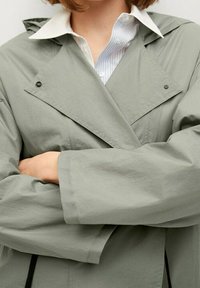 Lichtgroene, lichtgewicht jas met een oversized kraag, drukknoopsluiting aan de voorkant en brede mouwen. Draagt over een blauw gestreept overhemd.