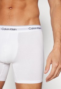 Boxeri scurți din bumbac alb, cu un logo negru Calvin Klein pe talie. Textură netedă, design ajustat și lungime până la mijlocul coapsei.