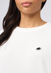 Weißer gerippter Pullover mit Rundhalsausschnitt und langen Ärmeln, ausgestattet mit einem kleinen schwarzen Mammut-Logo auf der Brust.