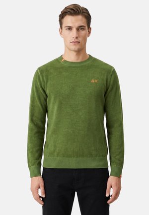 Uomo che indossa un maglione verde con scollo rotondo e piccolo logo arancione sul petto sinistro, abbinato a pantaloni neri, in piedi davanti a uno sfondo semplice.