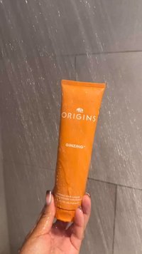 Oransje tube med Origins GinZing skrubb-rens med hvit tekst, våt overflate, holdt i en hånd, med glatt tekstur og avrundet form.