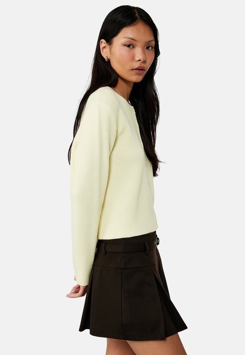 Femme aux longs cheveux noirs portant un pull côtelé crème et une jupe noire plissée, debout de profil sur un fond blanc.