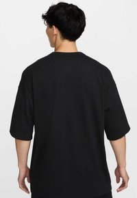 Vīrietis ar īsiem tumšiem matiem, tērpies vienkāršā melnā oversize T-kreklā, ir vērsts prom no kameras uz balta fona.