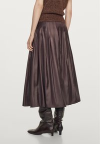 Lång, plisserad kjol i mörkbrun satin med en svängande silhuett. Kombinerad med åtsittande, mörka läderstövlar som går upp till knäna.