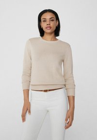 Beige gestrickter Pullover mit rundem Halsausschnitt und gerippten Bündchen, kombiniert mit weißen Hosen und einem braunen Gürtel. Einfaches, strukturiertes Design.