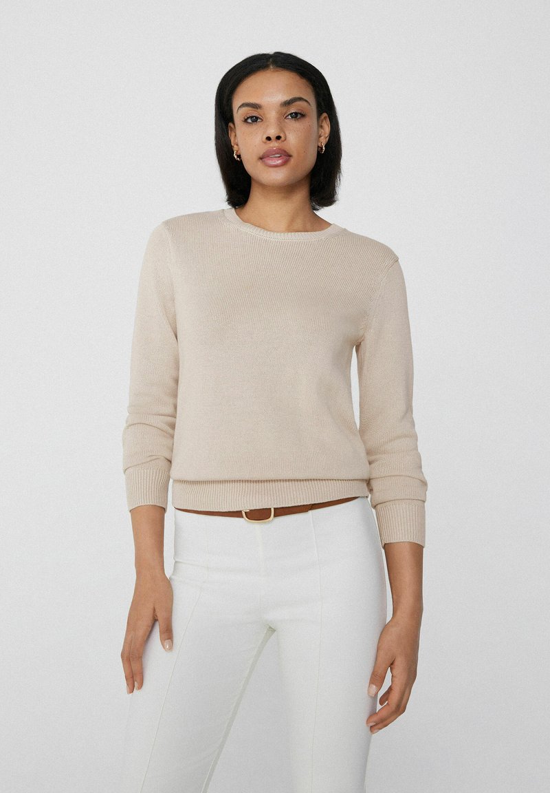 Beige gestrickter Pullover mit rundem Halsausschnitt und gerippten Bündchen, kombiniert mit weißen Hosen und einem braunen Gürtel. Einfaches, strukturiertes Design.