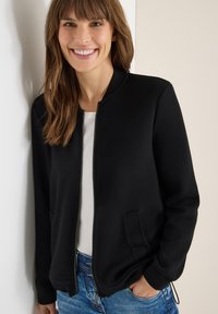 Chaqueta bomber negra con cremallera, de textura suave, cuello redondo y bolsillos laterales, combinada con una camisa blanca y jeans de mezclilla azul.