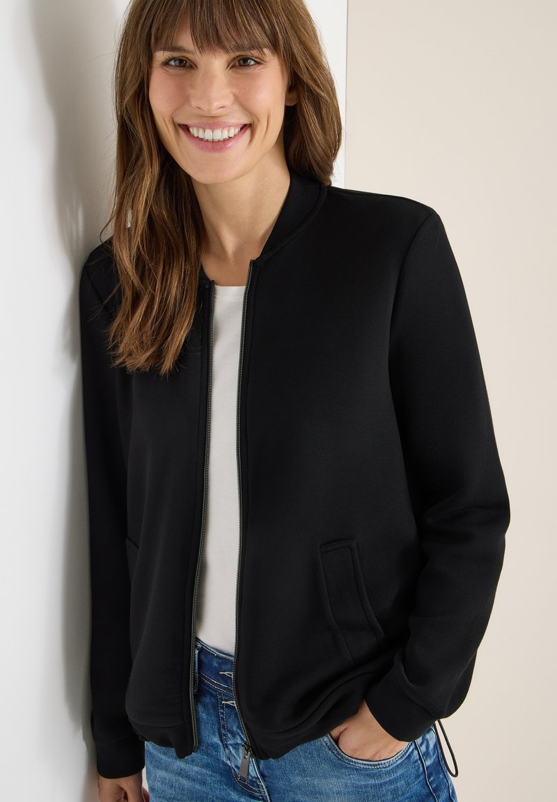 Chaqueta bomber negra con cremallera, de textura suave, cuello redondo y bolsillos laterales, combinada con una camisa blanca y jeans de mezclilla azul.