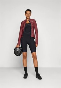 Cyklistický outfit zahrnuje bordó lehkou bundu, černé přiléhavé kalhoty na kšandy a odpovídající černou obuv. V jedné ruce je držen černá helma.