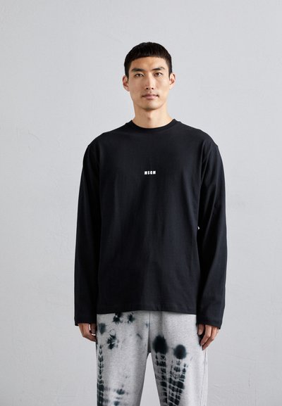 Cămașă neagră cu mânecă lungă, guler rotund și un logo alb "MSGN" centrat pe piept, asociată cu pantaloni de trening gri cu efect tie-dye, având modele circulare.