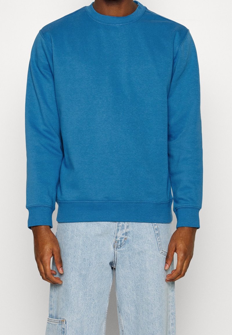 Personne portant un sweat-shirt à manches longues bleu uni et un jean denim bleu clair avec une poche sur la cuisse gauche, sur un fond blanc.