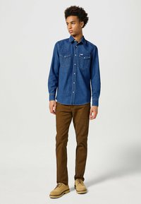 Camicia di denim blu scuro con bottoni a pressione, due tasche sul petto e cuciture in stile western. Abbinata a pantaloni marroni testurizzati e stivali color sabbia.