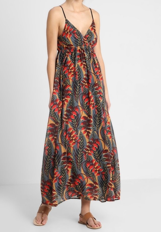 Femme portant une robe maxi sans manches avec un motif de feuilles tropicales rouge et vert foncé, associée à des sandales plates couleur beige.