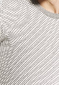 Zbliżenie na osobę w jasnoszarej, teksturowanej swetrze z subtelnym wzorem w kratkę oraz ściągaczem przy kołnierzu.